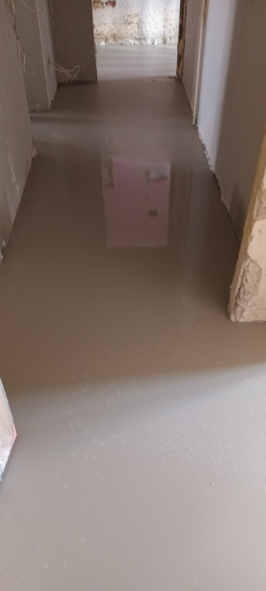 Wylany, mokry podkład podłogowy w trakcie prac remontowych. Widoczne surowe ściany i przejście do kolejnego pomieszczenia. Podłoże jest gładkie i jednolite.