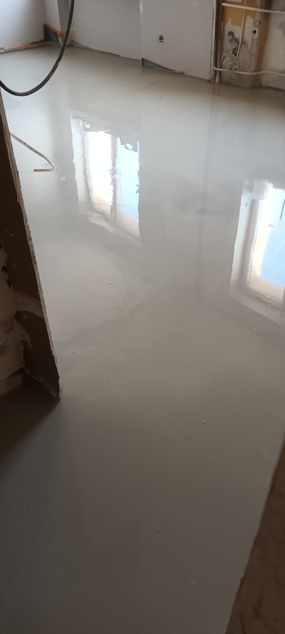 Świeżo wylana, gładka posadzka samopoziomująca w jasnym odcieniu szarości, odbijająca światło okien. Widoczne surowe ściany w trakcie prac wykończeniowych.