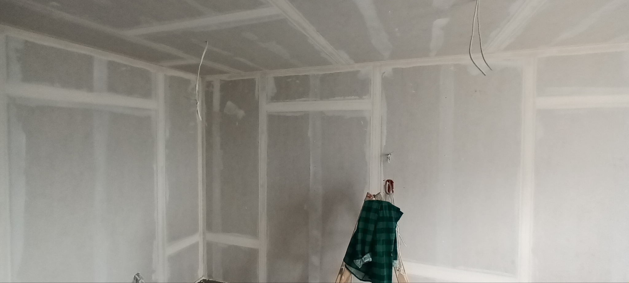 Wnętrze w trakcie prac wykończeniowych: ściany i sufit pokryte płytami gipsowo-kartonowymi, widoczne łączenia i szpachlowanie. Na drabinie leży zielona flanelowa koszula.