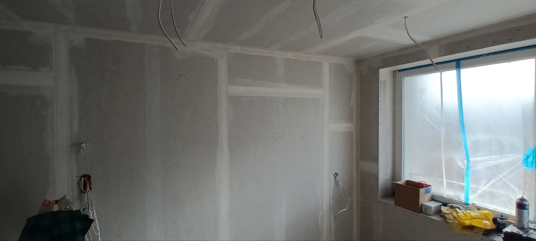 Pokój w trakcie prac wykończeniowych: ściany i sufit pokryte płytami gipsowo-kartonowymi, zabezpieczone okno folią malarską, widoczne kable elektryczne.