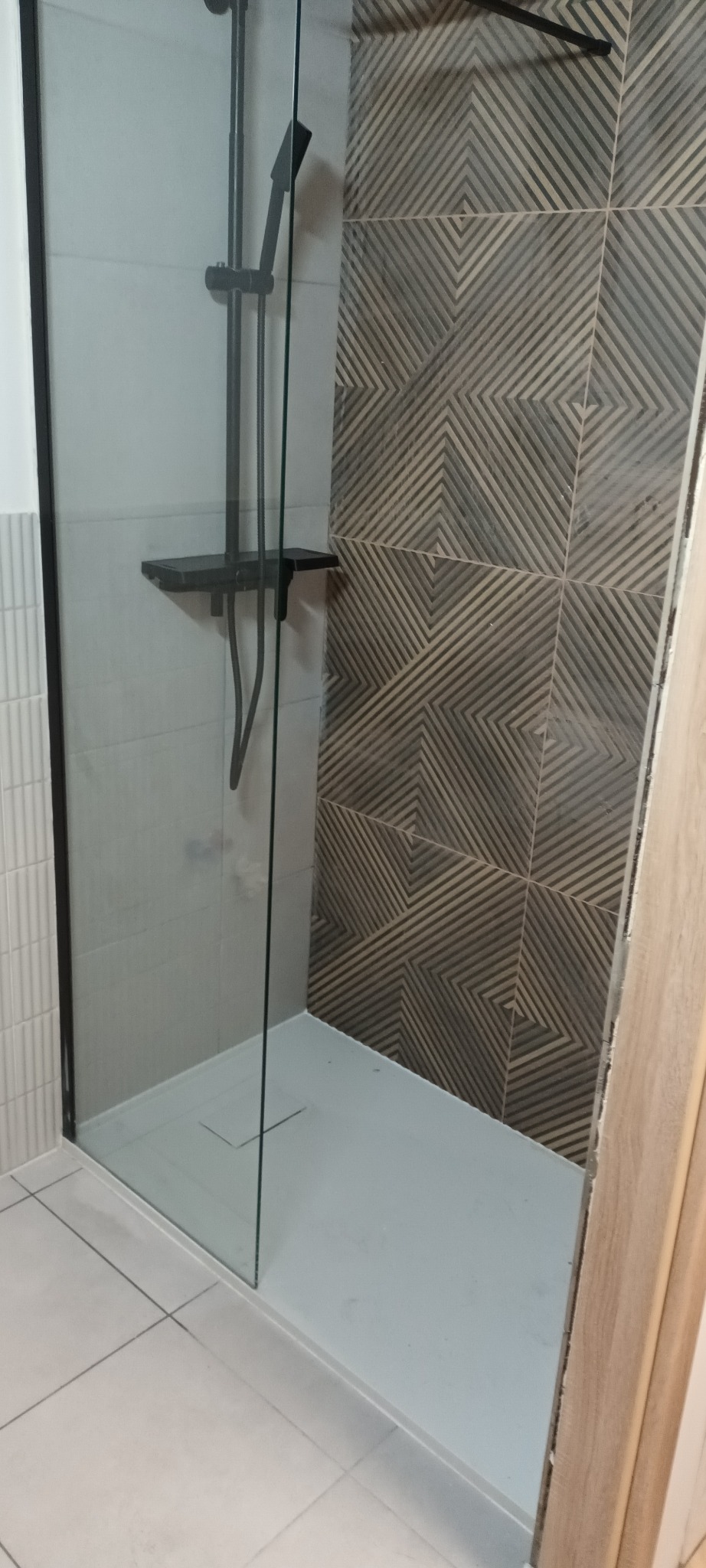 Nowoczesna kabina prysznicowa z czarną armaturą i geometrycznymi płytkami. Szklana ściana kabiny z czarną ramą. Minimalistyczny design, jasne płytki podłogowe.