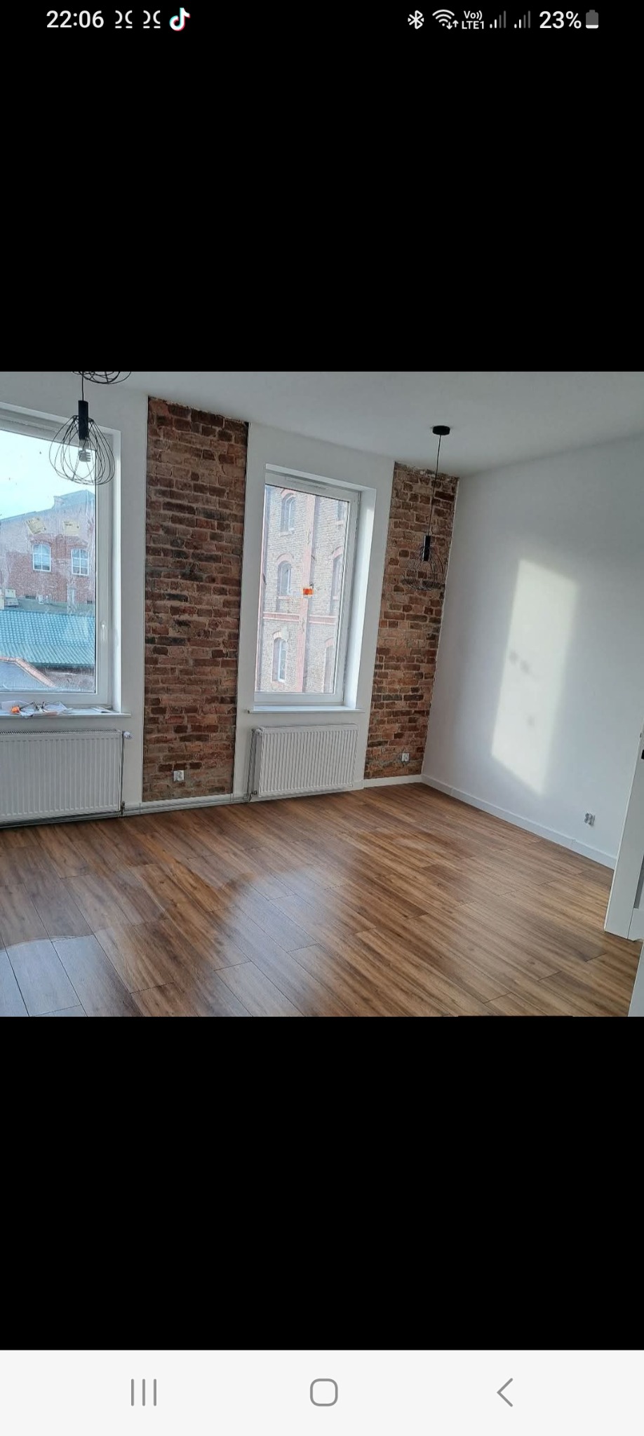 Jasne wnętrze po remoncie: drewniana podłoga, białe ściany z fragmentami cegły, okna z widokiem na budynki. Minimalistyczne lampy wiszące.