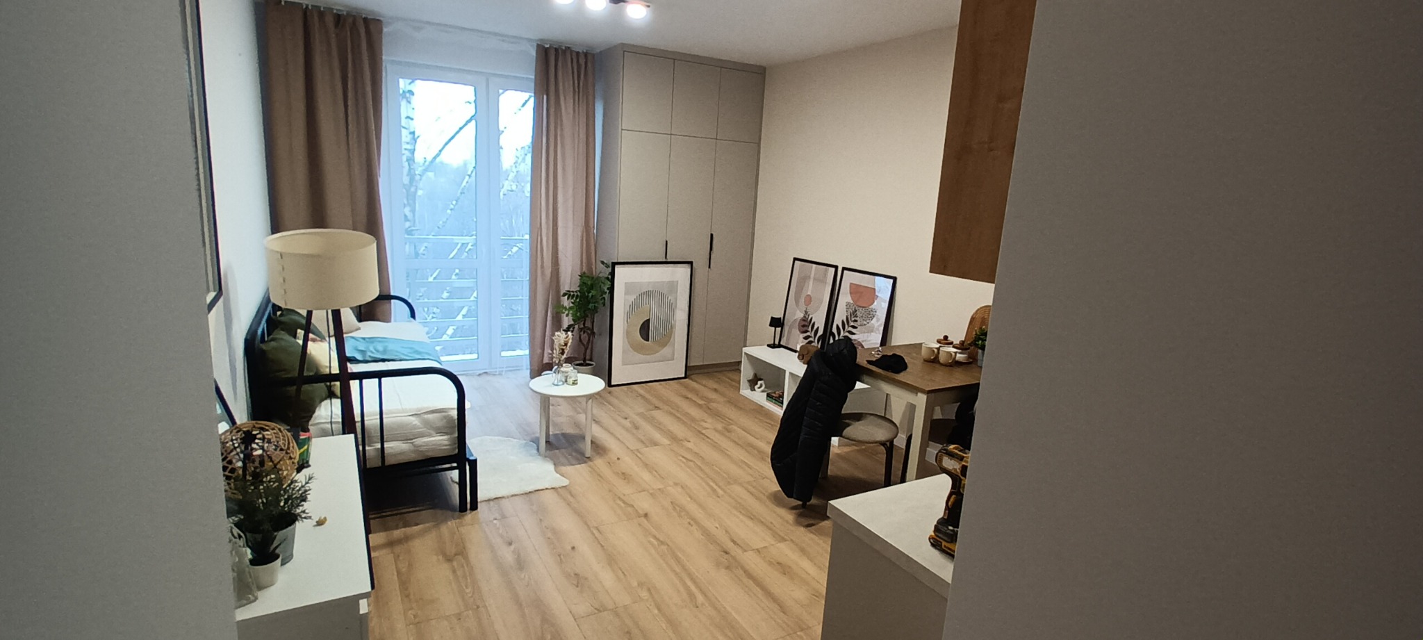 Jasne wnętrze pokoju z drewnianą podłogą, balkonem i minimalistycznym wystrojem. Widoczne łóżko, stół, szafa i dekoracje ścienne w neutralnych barwach.