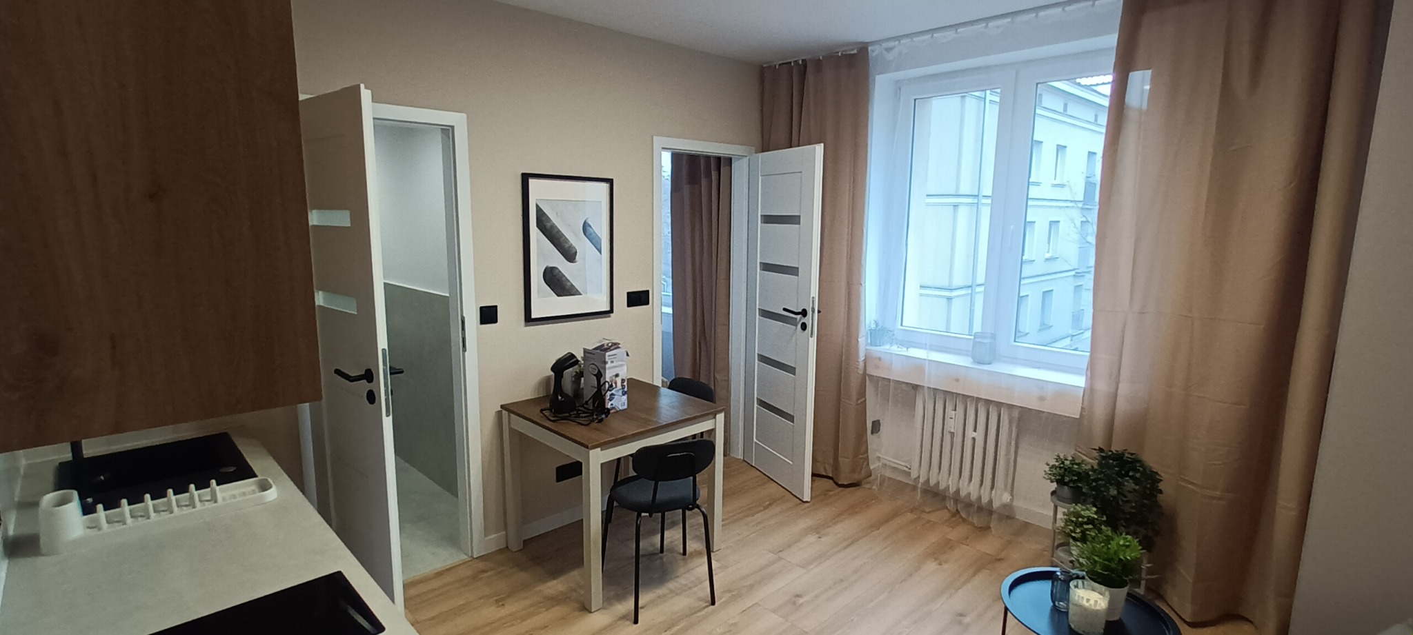 Jasne wnętrze mieszkania po remoncie: drewniana podłoga, stół z krzesłami, okno z beżowymi zasłonami, drzwi i dekoracje ścienne. Widok na odnowione pomieszczenie.