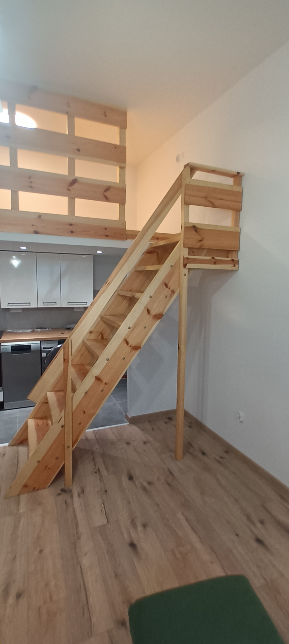 Drewniane schody prowadzące na antresolę z balustradą, widoczne na tle białej ściany i fragmentu kuchni. Naturalne drewno, prosta konstrukcja, jasne wnętrze.