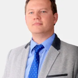 Mężczyzna w szarej marynarce, błękitnej koszuli i niebieskim krawacie w kropki, na jasnym tle. Portret agenta ubezpieczeniowego.