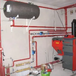 Złożona instalacja CO z piecem na paliwo stałe, zbiornikiem akumulacyjnym i licznymi rurami w kolorze czerwonym, zamontowana w kotłowni. Widoczne elementy sterujące i pompy obiegowe.