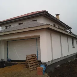 Dwukondygnacyjny dom z garażem w trakcie tynkowania elewacji, widoczne rusztowania i materiały budowlane. Częściowo zakryte okno garażowe folią.