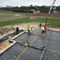 Wylewanie betonu na przygotowany fundament zbrojony siatką stalową, widoczni pracownicy w odblaskowych kamizelkach i pompa do betonu, w tle krajobraz z domami.