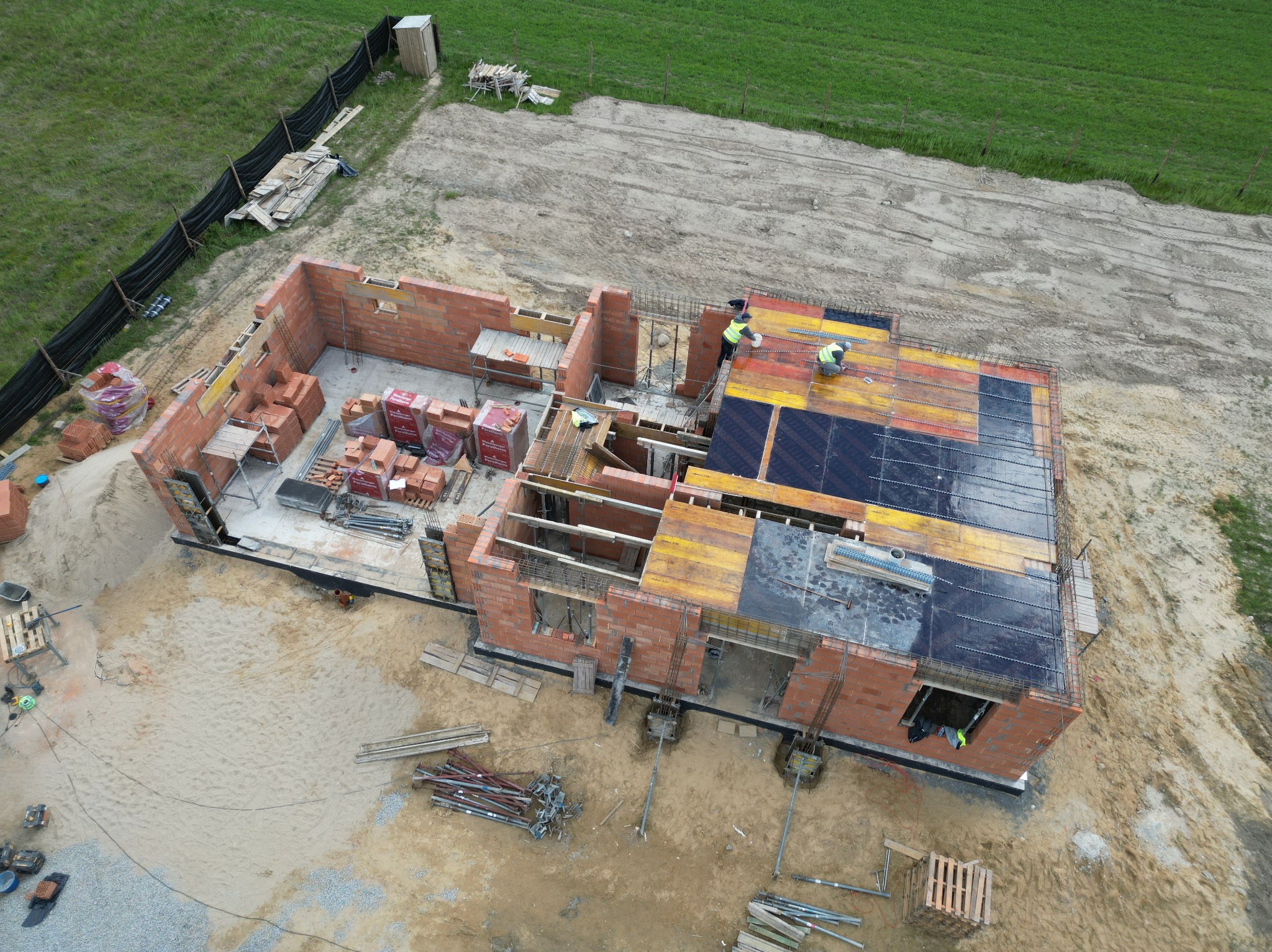Widok z lotu ptaka na plac budowy domu z czerwonej cegły, z widocznymi pracownikami w żółtych kamizelkach układającymi zbrojenie na przygotowanym szalunku stropu, paletami z cegłami i narzędziami...