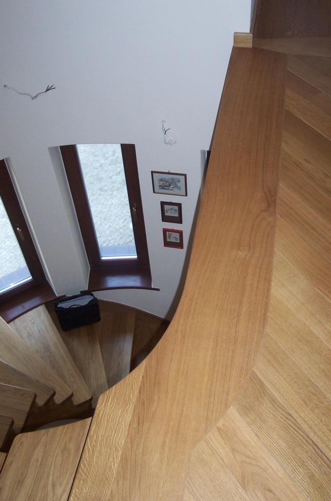 Kątowe ujęcie drewnianych schodów spiralnych z balustradą w naturalnym odcieniu drewna. Widoczne okna i dekoracje ścienne. Schody wykonane z jasnego drewna.