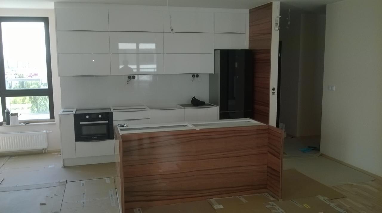 Nowoczesna kuchnia z białymi szafkami i drewnianym blatem wyspowym. Widoczne okno i czarna lodówka. Podłoga zabezpieczona kartonem na czas montażu.