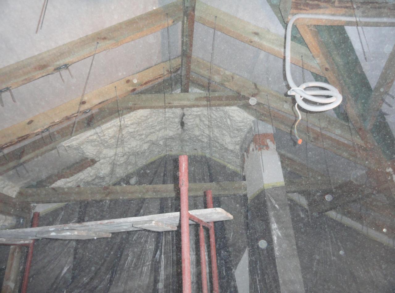 Poddasze ocieplone pianką, widoczne drewniane elementy konstrukcyjne dachu, rusztowanie i wystający komin, zabezpieczone folią. Widoczne wiszące przewody elektryczne.