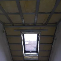 Poddasze z oknem dachowym w trakcie izolacji, widoczna wełna mineralna pomiędzy metalowymi profilami konstrukcyjnymi.