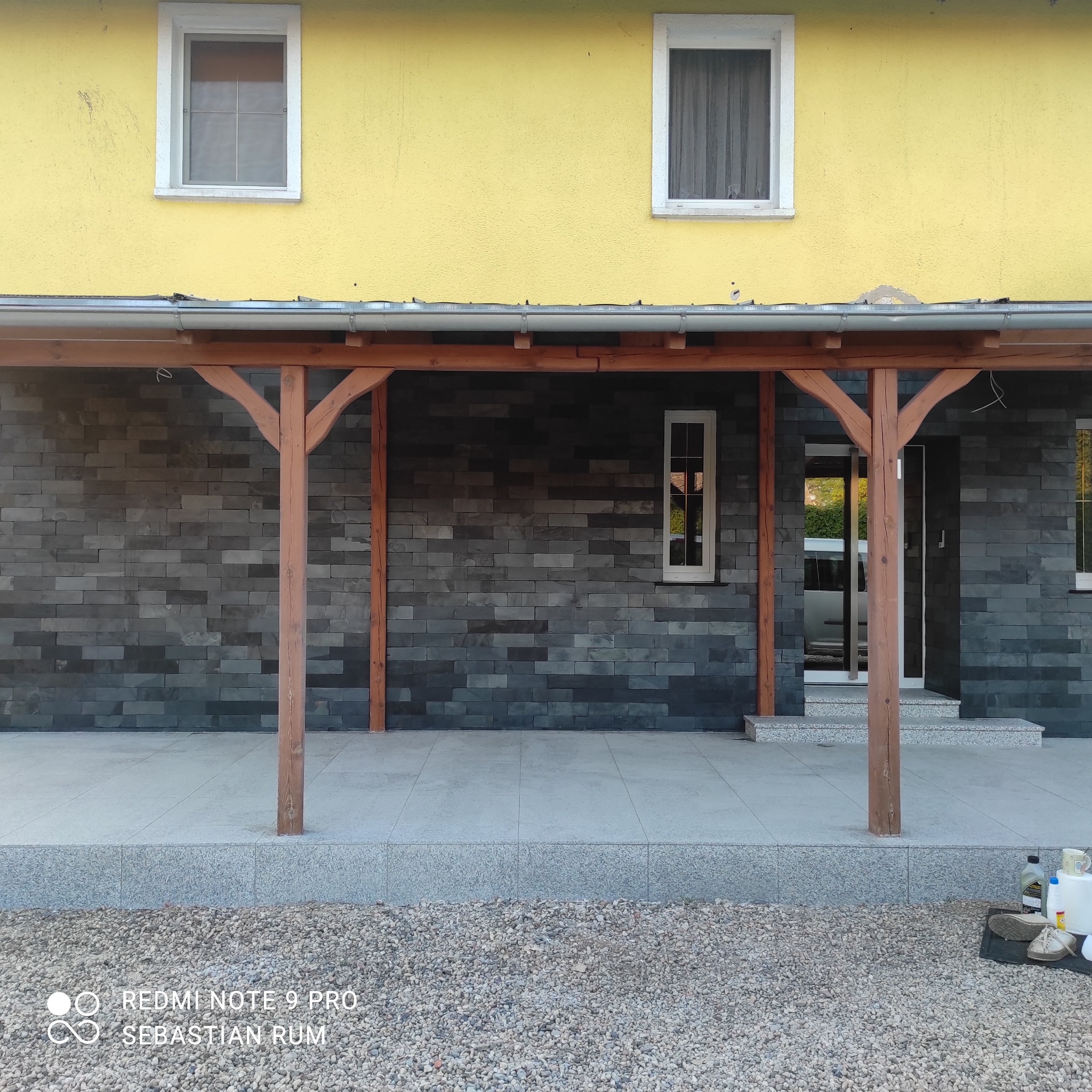 Elewacja domu z szarej cegły, drewniana konstrukcja zadaszenia tarasu, żółta ściana budynku z dwoma oknami. Widoczne płytki na tarasie i kamienie ozdobne.