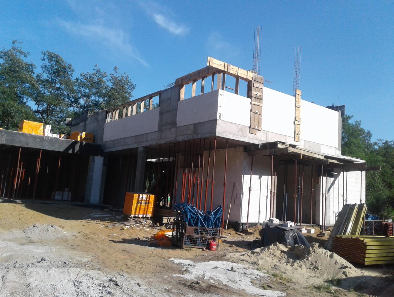 Budowa domu z betonu komórkowego w stanie surowym otwartym, widoczne podpory szalunkowe i zbrojenie, teren budowy z piaskiem i materiałami budowlanymi.