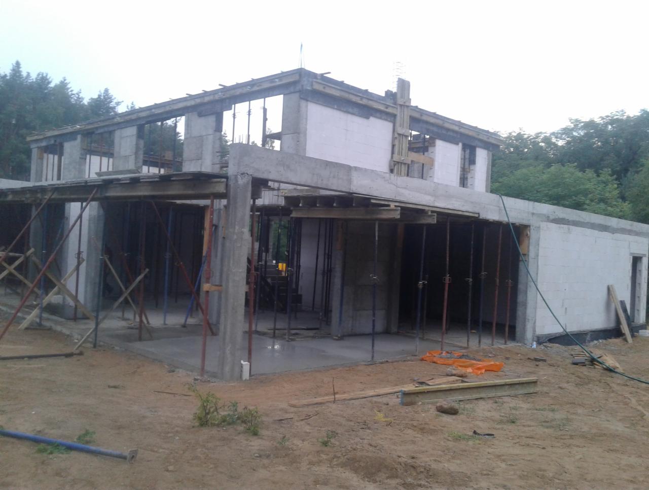 Dwukondygnacyjny dom w trakcie budowy, widoczne ściany z białych bloczków, betonowe słupy podtrzymujące strop, drewniane szalunki i metalowe podpory, teren wokół budynku jeszcze niezagospodarowany.