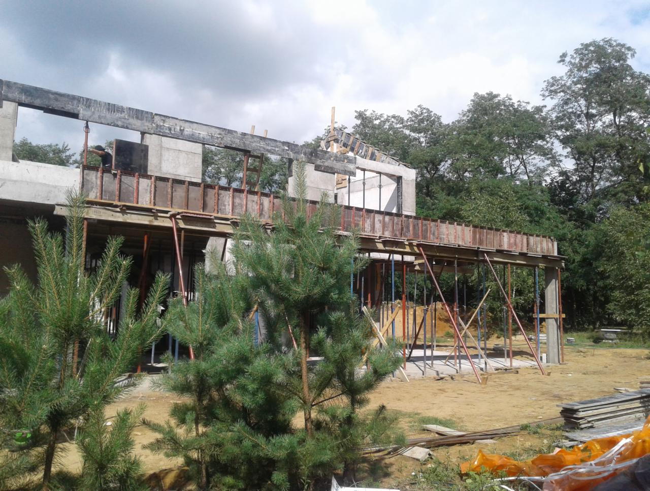 Surowa konstrukcja dwukondygnacyjnego budynku w trakcie budowy, z widocznym szalunkiem i podporami, otoczona zielenią i młodymi sosnami.