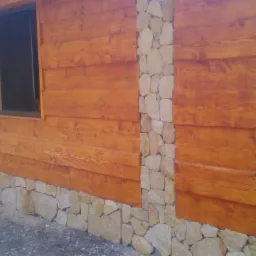 Drewniana elewacja budynku z oknem, wykończona kamiennymi elementami dekoracyjnymi. Jasne kamienie tworzą kontrast z ciepłym odcieniem drewna.