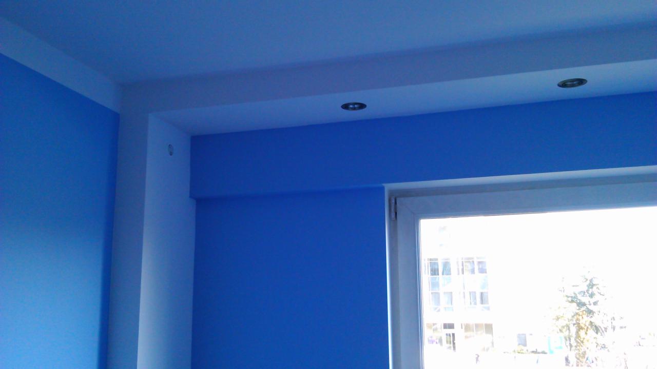 Niebieskie ściany po malowaniu z białymi listwami i oknem. Widoczne oświetlenie sufitowe. Jasne, naturalne światło wpada przez okno.