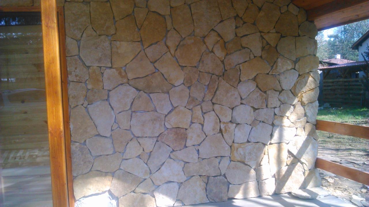 Ściana wykończona nieregularnymi, jasnymi kamieniami, widoczna drewniana konstrukcja i fragment otoczenia z budynkiem w tle. Naturalny kamień elewacyjny.