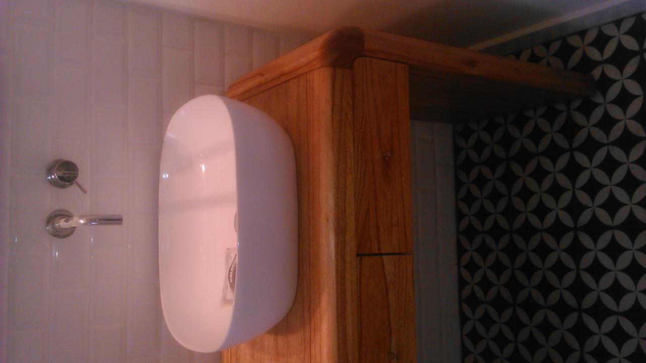 Wykończona łazienka: białe płytki, drewniana szafka z umywalką i bateria. Na ścianie wzorzysta tapeta. Widok z boku, ciepłe oświetlenie.