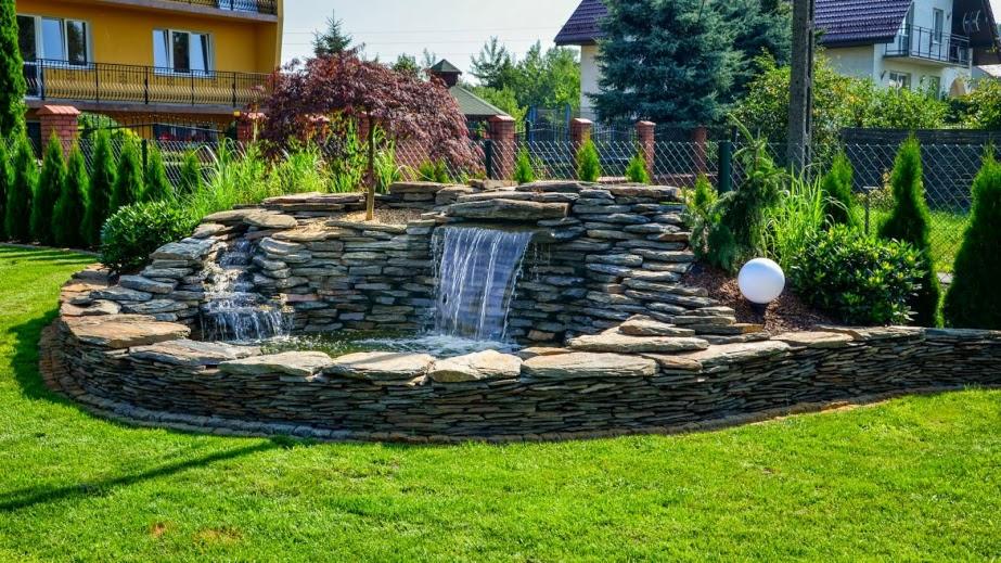 Kaskada wodna wykonana z płaskich kamieni łupkowych, otoczona zielonym trawnikiem i ozdobnymi krzewami, z kulistą lampą ogrodową po prawej stronie.