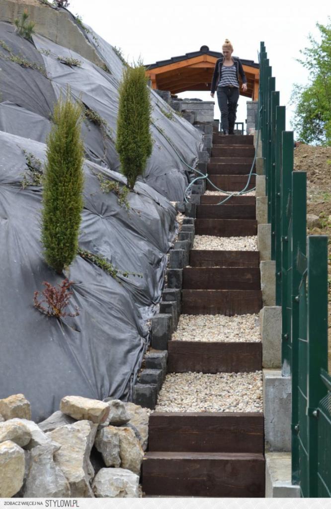Schody z drewnianymi stopniami i kamiennym wypełnieniem na zboczu zabezpieczonym czarną folią, prowadzące do drewnianej altany. Osoba schodzi po schodach.