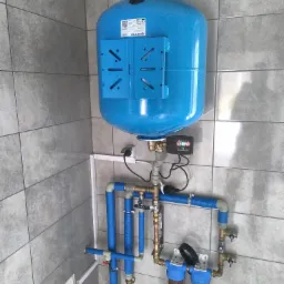 Niebieski zbiornik hydroforowy Aquasystem z osprzętem, filtrami wody i rurami w izolacji, zamontowany na szarej, kafelkowanej ścianie w pomieszczeniu.