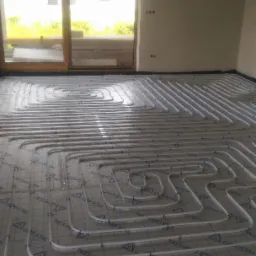 Białe rury ogrzewania podłogowego ułożone spiralnie na folii z nadrukiem GOR-STAL, widok z góry, okno balkonowe w tle, data 08.02.2018 w prawym dolnym rogu.