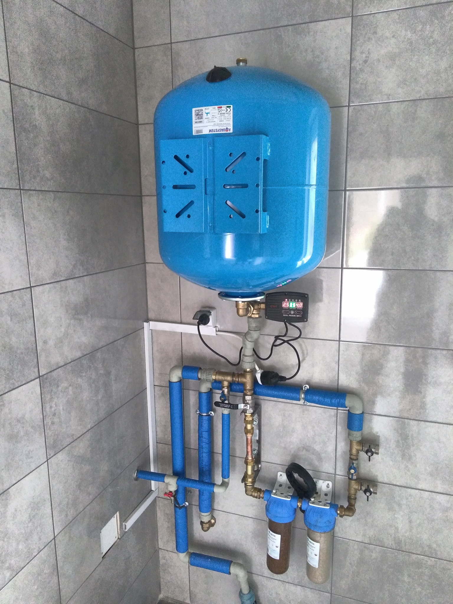 Niebieski zbiornik hydroforowy Aquasystem z osprzętem, filtrami wody i rurami w izolacji, zamontowany na szarej, kafelkowanej ścianie w pomieszczeniu.