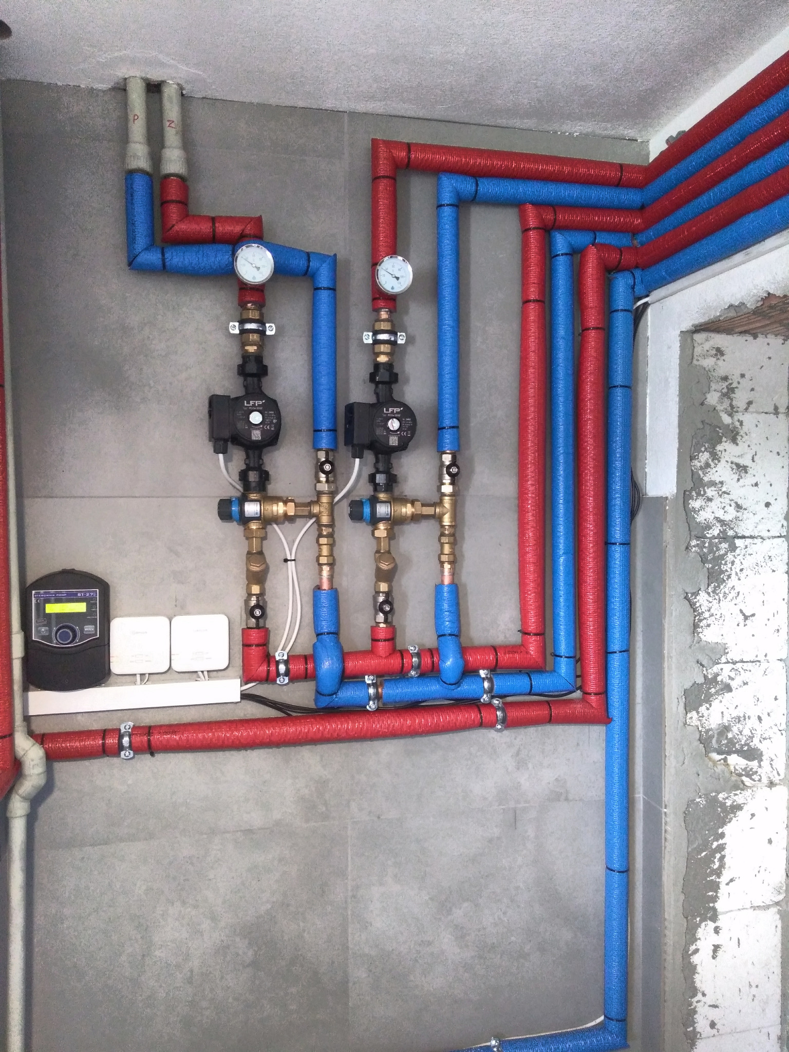 Instalacja grzewcza z rurami w izolacji czerwonej i niebieskiej, manometry, pompy obiegowe i regulator temperatury zamontowane na szarej ścianie w trakcie montażu.