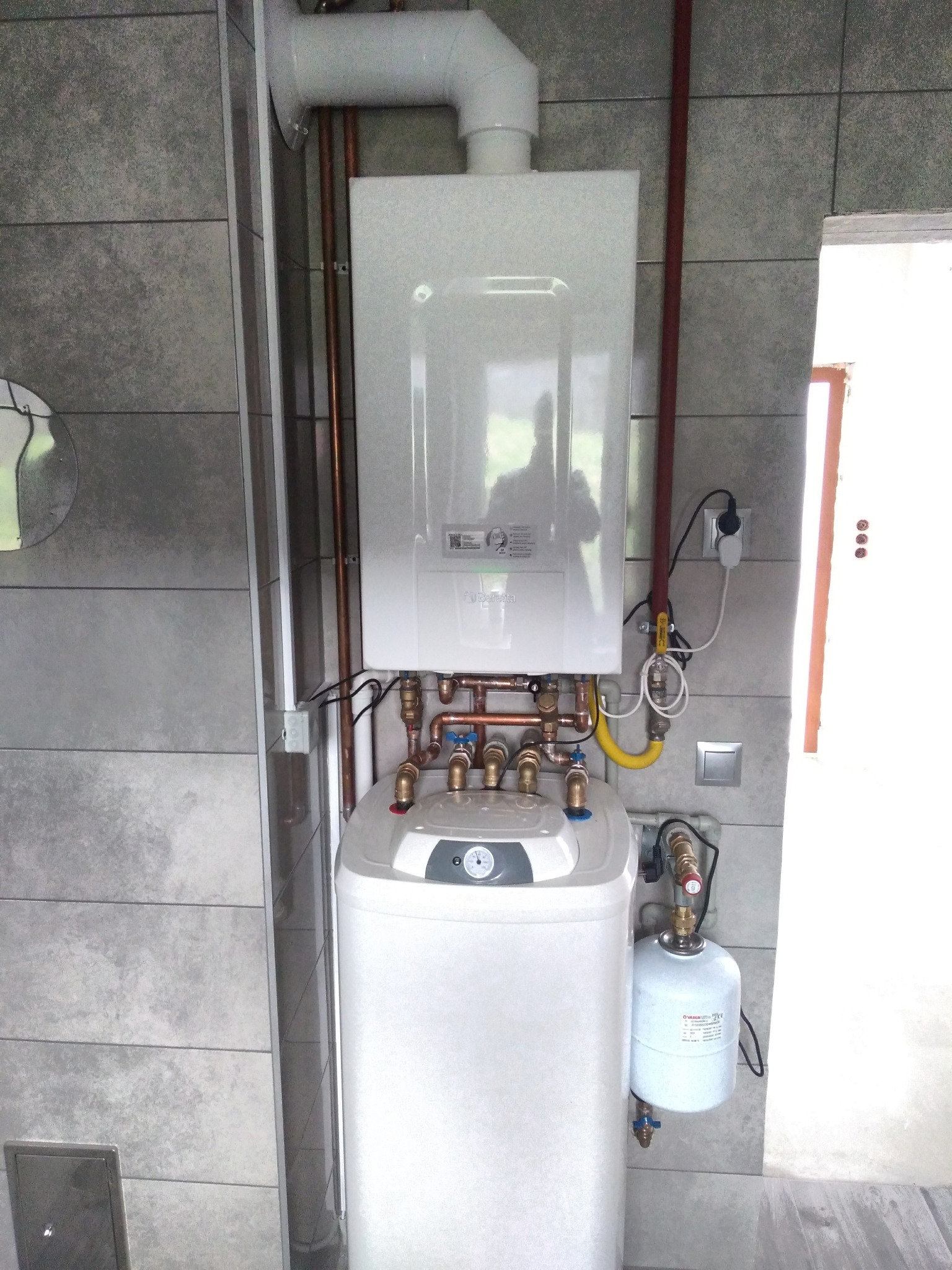 Nowoczesny, biały piec gazowy Beretta z zasobnikiem, zamontowany na szarej, kafelkowanej ścianie, widoczne miedziane rury i żółty przewód gazowy, podłączony do gniazdka elektrycznego, odbicie...