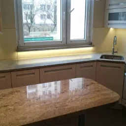 Jasny blat kamienny w kuchni z oknem, podświetlony ledami, z widocznym zlewem i płytą grzewczą. Na pierwszym planie fragment blatu stołu z odbiciem.