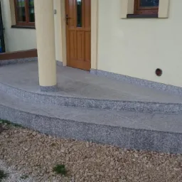 Wejście do domu z dwustopniowymi schodami pokrytymi szarym, nakrapianym kamieniem, z kolumnami podtrzymującymi zadaszenie i brązowymi drzwiami wejściowymi.