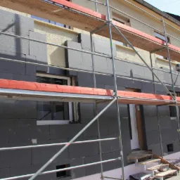 Ocieplanie elewacji budynku jednorodzinnego styropianem grafitowym, widoczne rusztowanie i fragmenty nieocieplonej ściany.