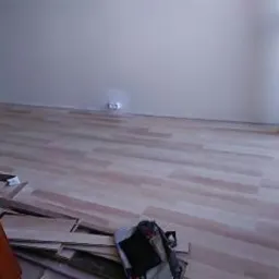 Układanie paneli podłogowych w jasnym pokoju, widoczne niedocięte panele i torba narzędziowa na podłodze, ściany pomalowane na jasny kolor, gniazdko elektryczne na ścianie.