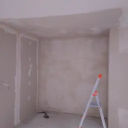 Wnętrze pokoju w trakcie prac wykończeniowych, widoczne płyty gipsowo-kartonowe na ścianach i sufitach, częściowo pokryte świeżą warstwą szpachli, aluminiowa drabina rozstawiona w rogu...