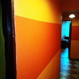 Firma Budowlano - Remontowa - Korytarz z pomalowanymi ścianami w poziome pasy w odcieniach żółtego i pomarańczowego, widoczne drzwi z zasłoną po lewej, oraz fragment podłogi w szachownicę.