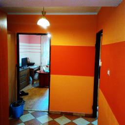 Firma Budowlano - Remontowa - Pomarańczowy korytarz z dwukolorowymi pasami na ścianie, widok na pokój z biurkiem i komputerem, podłoga w brązowo-białą szachownicę, niebieskie wiadro przy drzwiach.