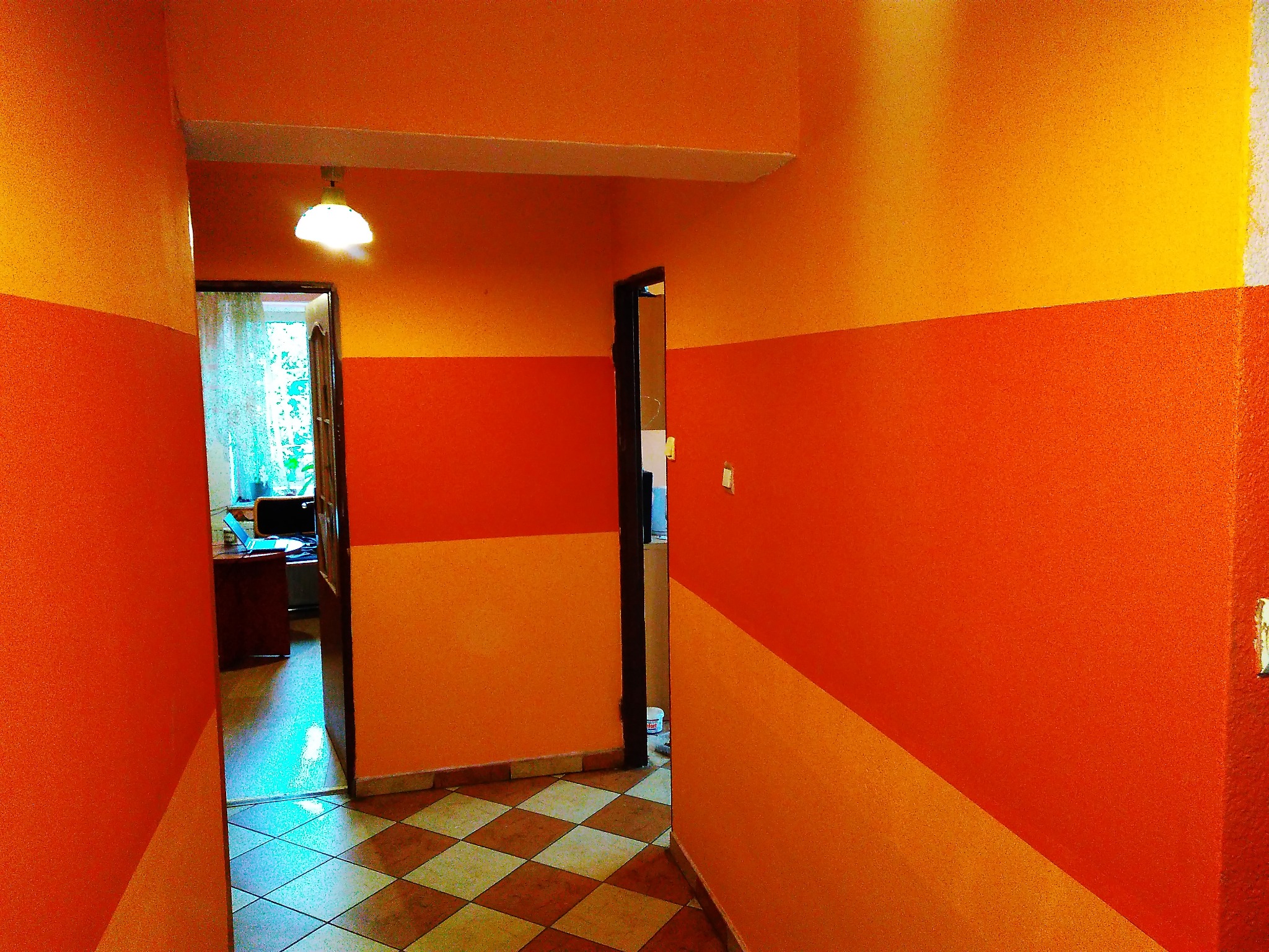 Korytarz z pomalowanymi ścianami w pasy w odcieniach pomarańczy i żółci, widoczne drzwi i podłoga w szachownicę.