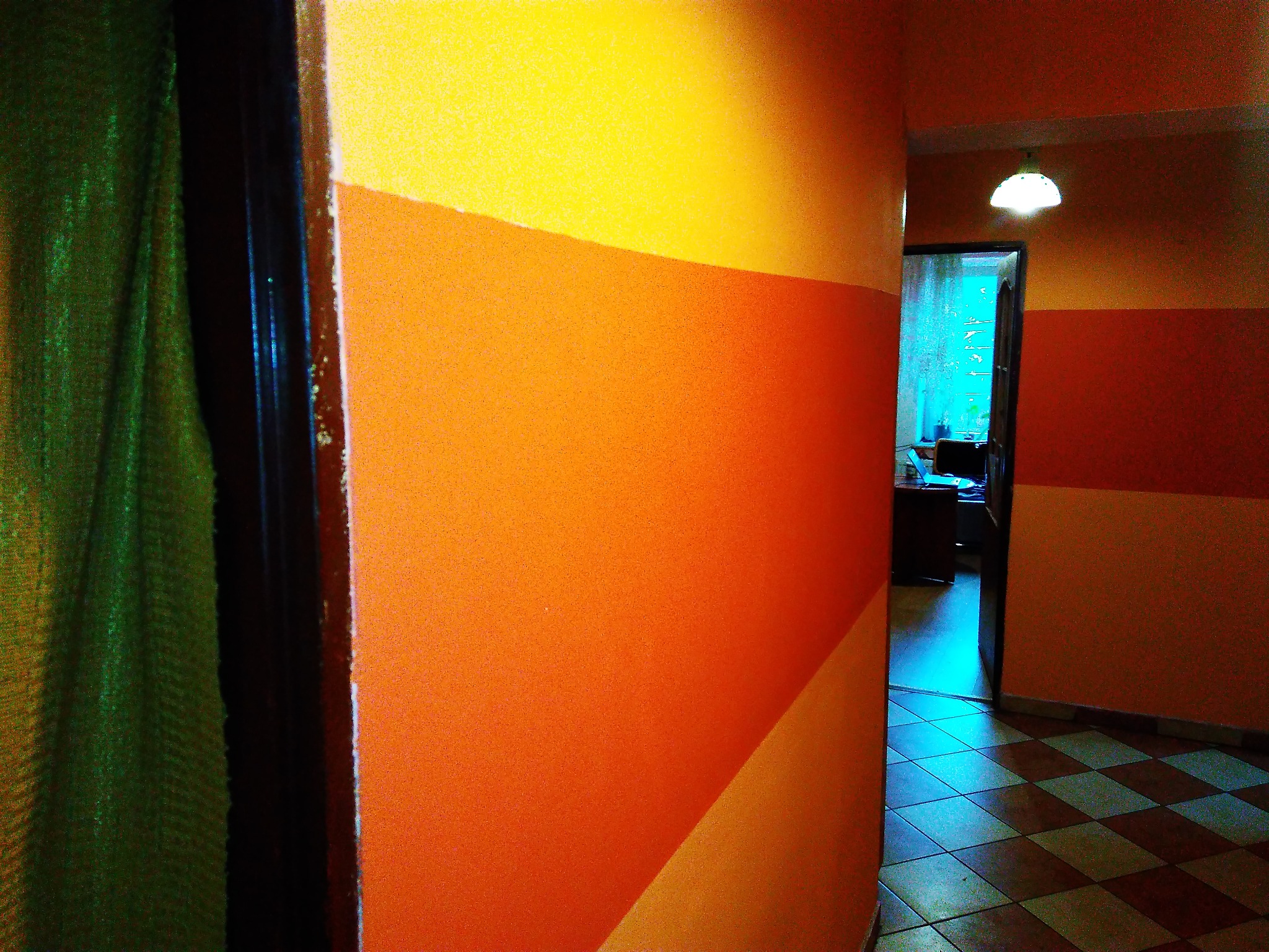 Korytarz z pomalowanymi ścianami w poziome pasy w odcieniach żółtego i pomarańczowego, widoczne drzwi z zasłoną po lewej, oraz fragment podłogi w szachownicę.