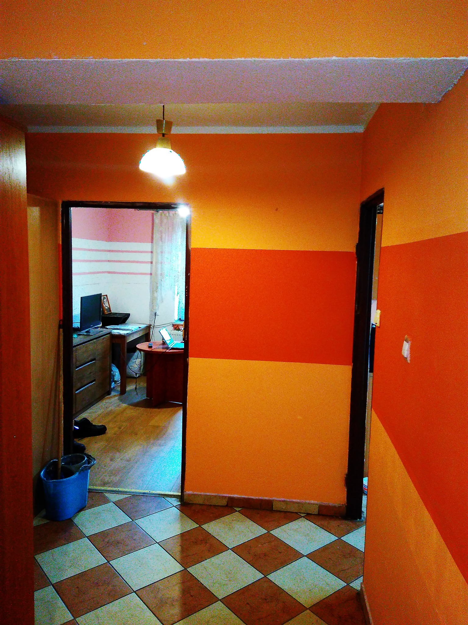 Pomarańczowy korytarz z dwukolorowymi pasami na ścianie, widok na pokój z biurkiem i komputerem, podłoga w brązowo-białą szachownicę, niebieskie wiadro przy drzwiach.