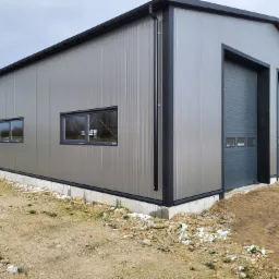 Nowoczesna hala magazynowa z szarych płyt warstwowych z dwoma bramami segmentowymi i oknami, widok z zewnątrz na nierówny teren z kamieniami.