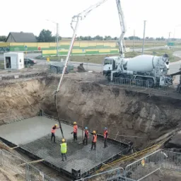 Wylewanie betonu na przygotowaną zbrojoną płytę fundamentową w wykopie, widoczne dwie betoniarki i pracownicy w kaskach i kamizelkach odblaskowych, ramię pompy do betonu doprowadza mieszankę.