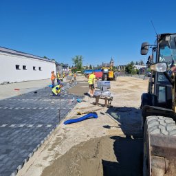 Andezyt - Brukarstwo, Kamieniarstwo - Pracownicy w odblaskowych kamizelkach układają szarą kostkę brukową na przygotowanym podłożu; w tle budynek, niebo bezchmurne, oraz ciężki sprzęt budowlany