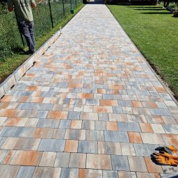 Andezyt - Brukarstwo, Kamieniarstwo - Długa, nowo ułożona ścieżka z kostki brukowej w odcieniach szarości i pomarańczy prowadząca przez trawnik w kierunku domu, widoczne rękawice robocze na kostce i pracownik przy ogrodzeniu.