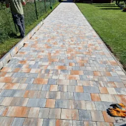 Długa, nowo ułożona ścieżka z kostki brukowej w odcieniach szarości i pomarańczy prowadząca przez trawnik w kierunku domu, widoczne rękawice robocze na kostce i pracownik przy ogrodzeniu.