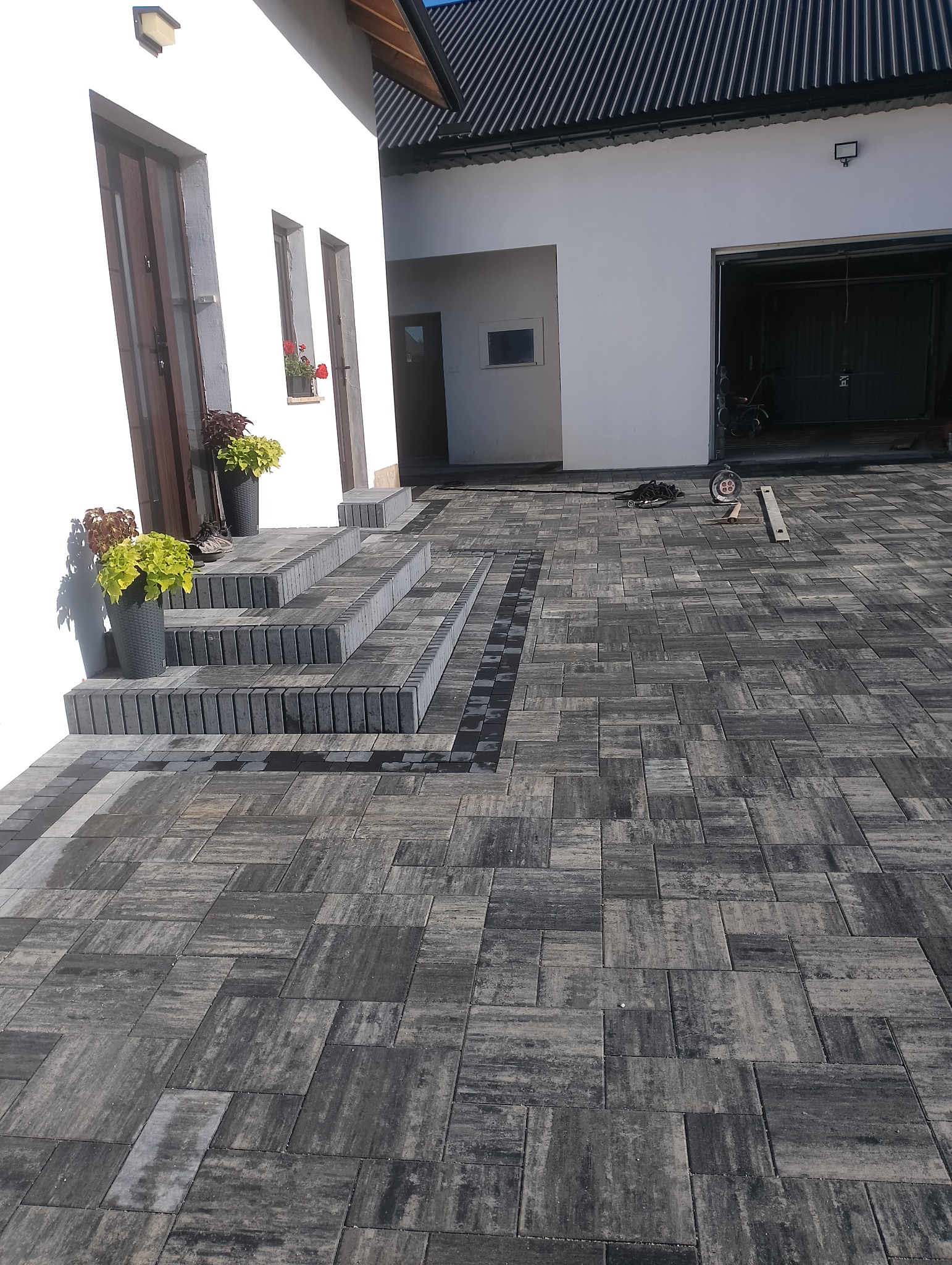 Nowoczesny podjazd z szarej kostki brukowej ułożonej w geometryczny wzór, z widocznymi schodami wejściowymi i elementami dekoracyjnymi w postaci donic z roślinami.
