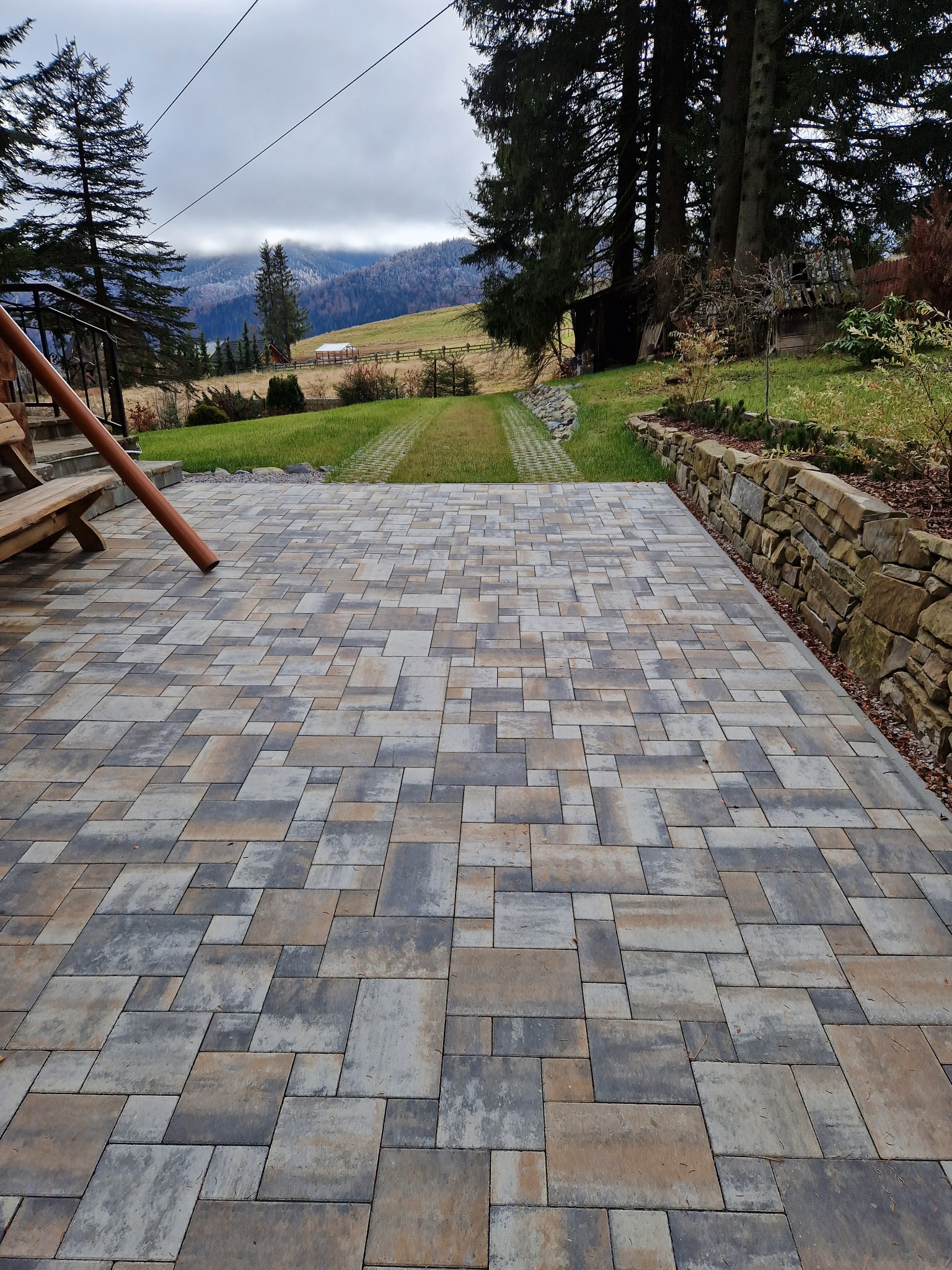 Nowo ułożona, wielobarwna kostka brukowa na posesji z widokiem na góry, otoczona trawnikiem i kamiennym murkiem oporowym.
